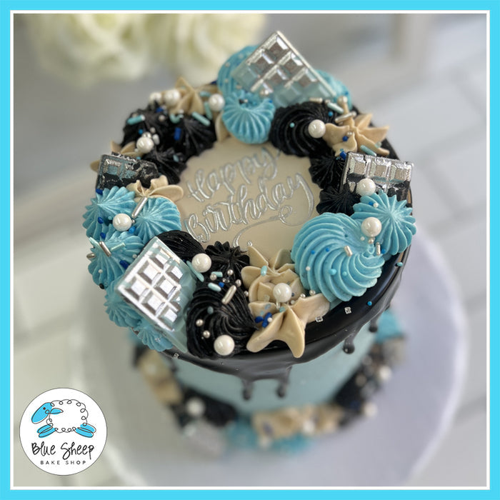 Black and blue drip birthday cake with metallic silver accents, buttercream swirls, and a custom "Happy Birthday" topper by Blue Sheep Bake Shop.