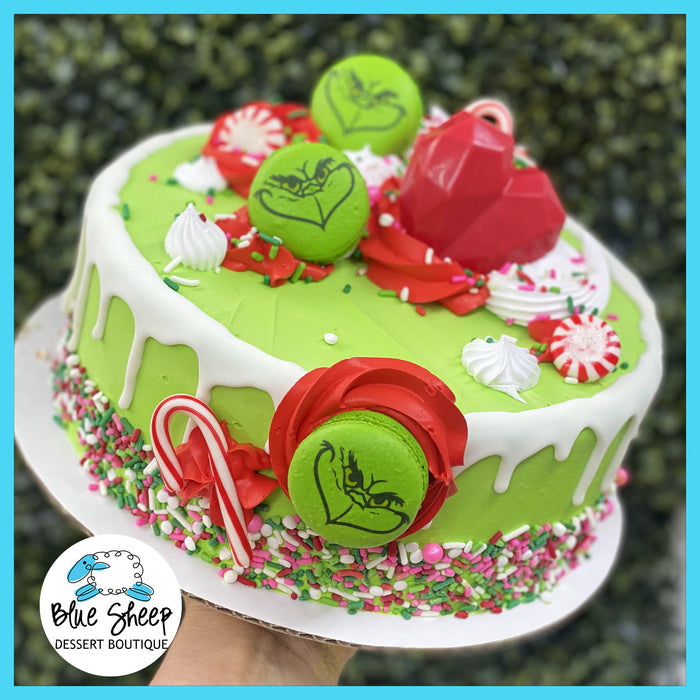 Grinch-themed ice cream cake with green frosting, white drip detail, Grinch macarons, candy canes, red chocolate heart, peppermint swirls, and festive sprinkles.