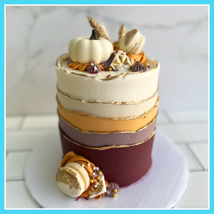 Thanksgiving-themed cake decorated in fall colors with gold leaf accents, topped with white chocolate pumpkins, meringue kisses, macarons, and fall sprinkles.