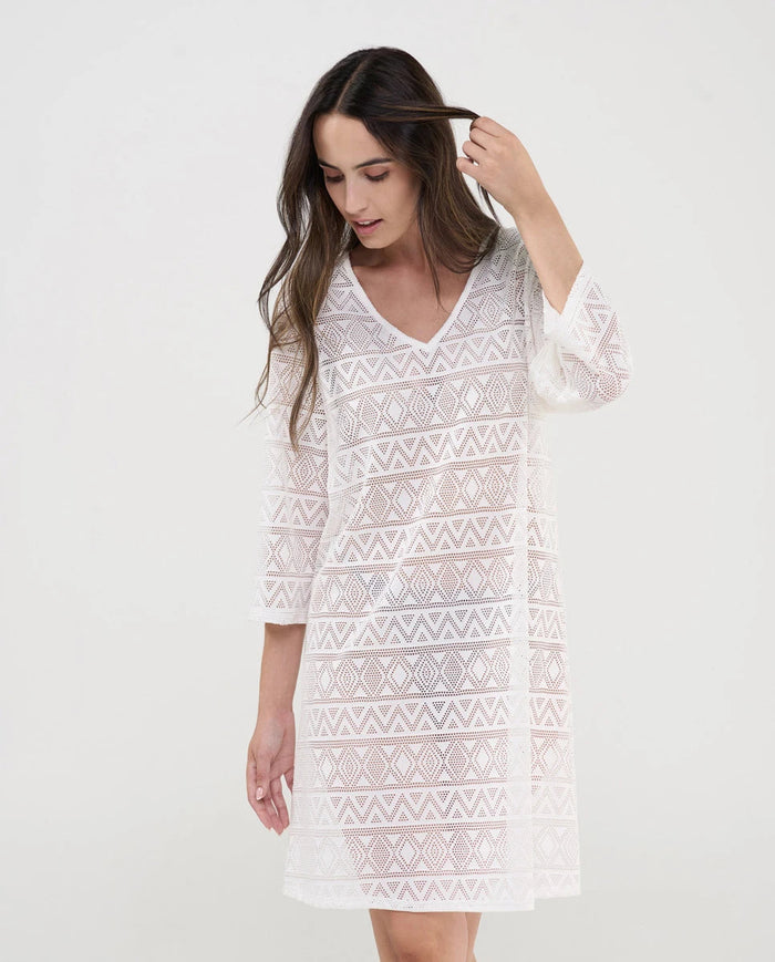 Mujer luce vestido corto blanco de baño con manga en punto desagujado motivos geométricos con cuello pico.