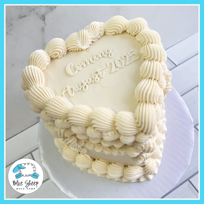 Heart-shaped baby announcement cake with piped vintage buttercream, elegant white frosting, and a custom message, available at Blue Sheep Bake Shop in Somerville, NJ